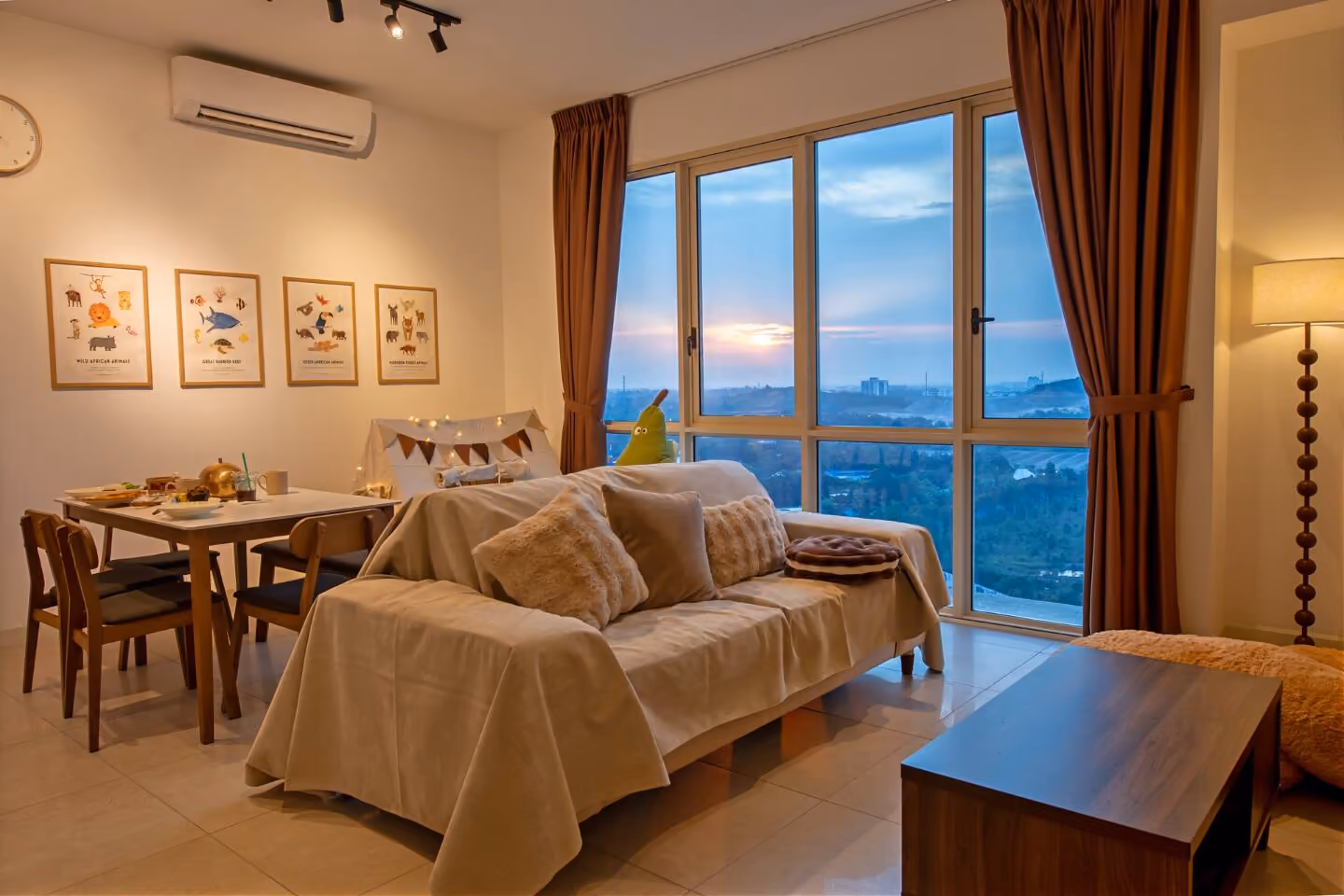The living room and dining area of a Medini apartment let by the night, with a sofa under a beige throw, a dining table laid for breakfast, framed animal prints on the wall, and floor-to-ceiling windows showing a sunset over Iskandar Puteri.