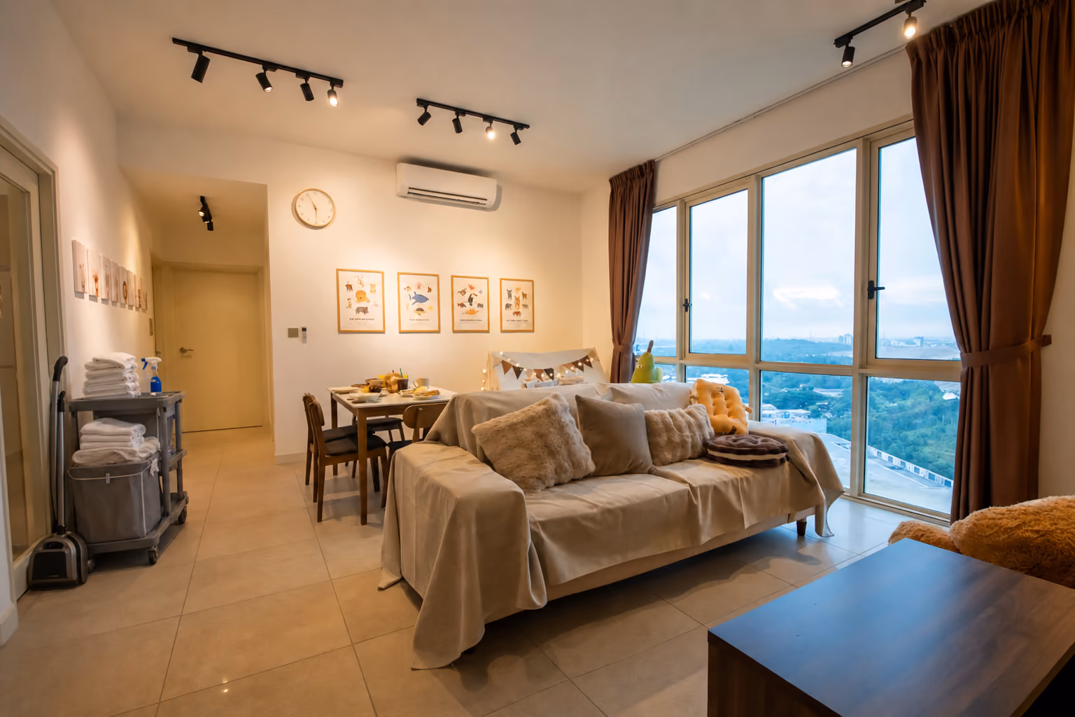 The hallway of a Medini apartment let by the night, with a housekeeping trolley parked by the front door, stacked with folded white towels, a basket of used linen and a vacuum.