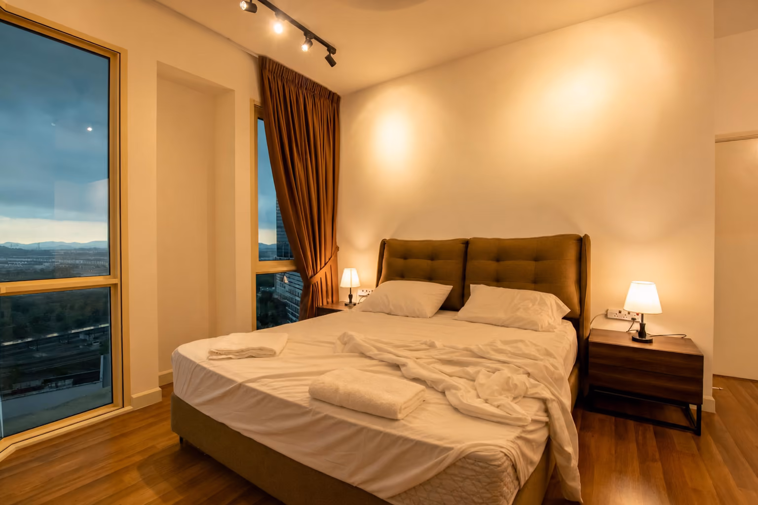 A bedroom in a Medini apartment between guests, the duvet stripped off, the pillows bare, the sheet pulled back across the mattress and two folded towels waiting on the bed.